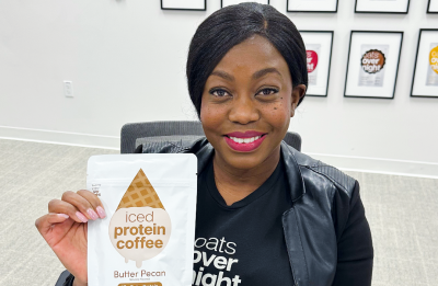 Person seated at a table holds a package labeled “Iced Protein Coffee, Butter Pecan,” with framed product posters displayed on the wall behind.