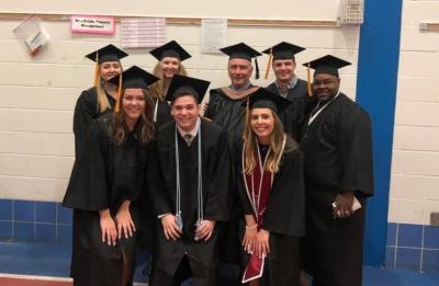 Group of eight people in graduation caps and gowns posing together in a gymnasium, standing in front of a tan wall with blue trim.