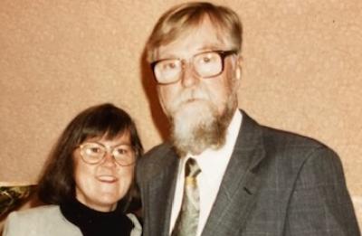  Two people stand together for a portrait, both wearing glasses, in an indoor setting.