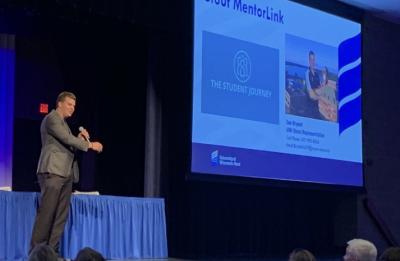 Speaker presents on stage with a slide reading “Stout MentorLink” to an audience seated in a lecture hall.