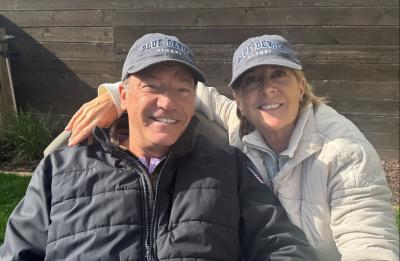 Two adults wearing “Blue Devils Alumni” hats sit outdoors on a bench, leaning together for a close-up photo with a wooden fence and greenery behind them.