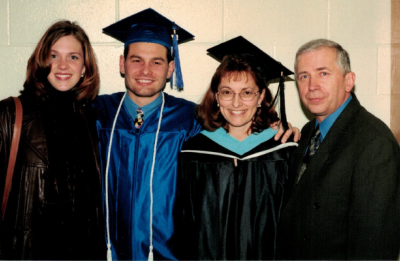 Four people posing indoors, including one individual wearing a cap and gown, with others standing close together against a plain wall.