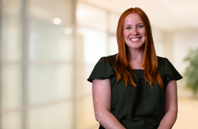 Person with long red hair smiling at the camera, seated against a softly blurred indoor background.