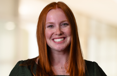 Person with long red hair smiling at the camera, seated against a softly blurred indoor background.