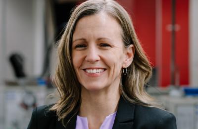 Person in a black blazer and light purple blouse, smiling at the camera, standing in an indoor lab or workspace with blurred equipment and red storage cabinets in the background.