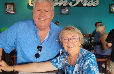 Two older adults seated at a restaurant table, smiling at the camera, with a turquoise wall and “Green Tree” sign behind them.
