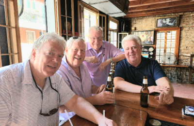 Four adults gathered around a wooden table inside a bar, with drinks in hand and exposed brick and wood interior in the background.