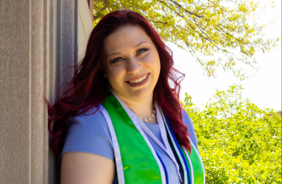 UW-Stout graduate posing outdoors while holding a diploma and wearing graduation cords, a medallion and a green stole.