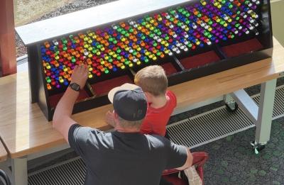 An adult and child interact with a colorful light peg wall exhibit, placing translucent pegs into a large backlit display at a children's museum.