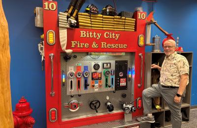 A person stands beside a large interactive "Bitty City Fire & Rescue" play structure at the Children’s Museum of Eau Claire. The fire truck-themed display features hoses, gauges, lights and firefighting equipment.