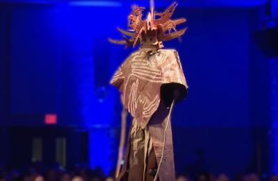 A student model walks a runway wearing an elaborate sculptural costume with a textured poncho and antler-like headpiece under blue stage lighting.