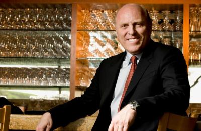 A person in a dark suit and red tie sits at a restaurant bar, with shelves of glassware displayed behind.