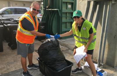 A person wearing a safety vest, gloves and a green cap helps sort waste materials into a large collection bin at an outdoor worksite.