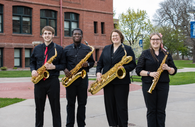 Four student musicians pose outdoors on the UW-Stout campus holding saxophones in front of a red brick academic building.