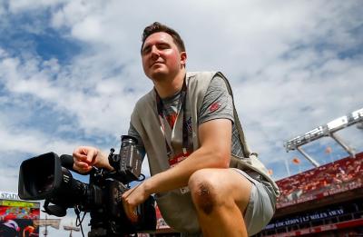 A camera operator kneels on the sideline of a football field beside a professional video camera before a game.