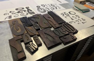 Large wooden letterpress type blocks in a variety of sizes and decorative styles are arranged on a metal worktable beside printed proofs showing matching letterforms. The collection highlights traditional typographic design and printmaking techniques.