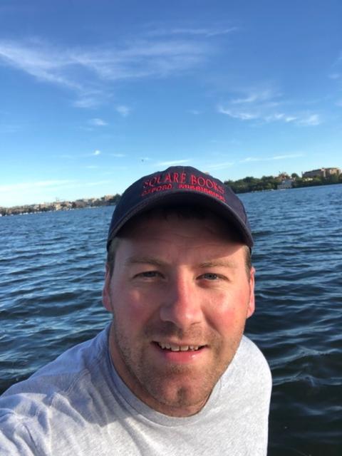 John Strauser, a white man wearing a baseball cap, smiles at the camera. There is a lake in the background
