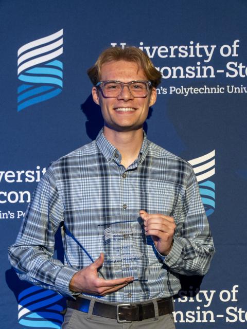 A person stands in front of an official University of Wisconsin–Stout backdrop while holding a clear award plaque. They are wearing a plaid shirt and slacks.