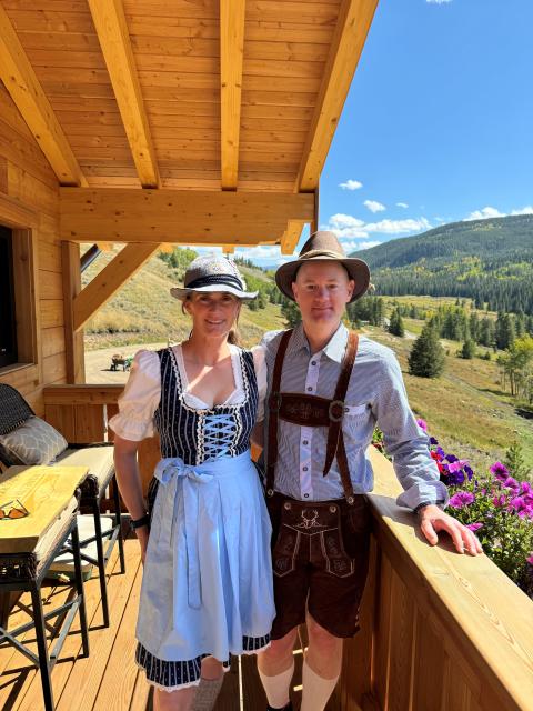 Two people wearing traditional Bavarian-style clothing standing on a wooden deck with views of rolling hills, wildflowers, and a sunny mountain landscape.