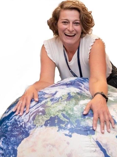 A person is leaning forward over a large inflatable globe, resting their hands on it while smiling toward the camera. They are wearing a sleeveless white top and a lanyard, with a watch on one wrist. The background is plain white.