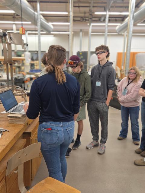 A student leads a small group of visiting students on a tour inside a woodshop classroom. The guide stands at a workbench with a laptop, while the group observes various tools, equipment, and workshop features around the space.