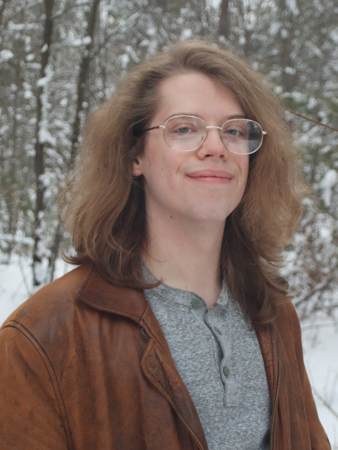 Outdoor winter portrait A person with long, wavy hair stands outside in a snowy forest, wearing glasses, a brown leather jacket, and a gray henley shirt.