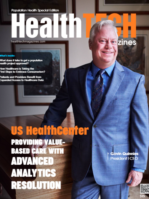 A person wearing a blue suit, blue dress shirt, and patterned tie stands with one hand resting on a wooden desk, featured on the cover of HealthTech Magazines. The headline highlights US HealthCenter and value‑based care supported by advanced analytics. Additional cover text previews articles inside the issue, and framed artwork is visible in the background.