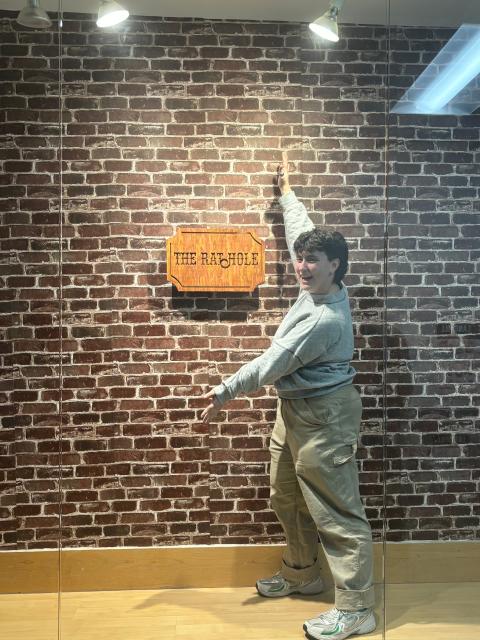 A person stands in front of a brick-patterned display wall, reaching upward with one arm. A wooden sign reading “THE RAT HOLE” is mounted on the wall under gallery-style lighting.
