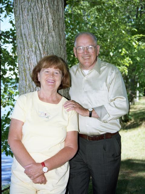 Two adults standing near a tree in a grassy, wooded area, facing the camera in casual clothing.