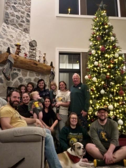 A large group of people gathers in a living room decorated for Christmas, seated and standing near a tall Christmas tree lit with white and red ornaments. A stone fireplace with holiday decorations and stockings hangs on the left, and a dog rests on the floor near the group.