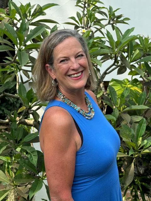 Person standing outdoors in front of leafy green plants, wearing a sleeveless blue top and a beaded necklace, with a white wall partially visible in the background.