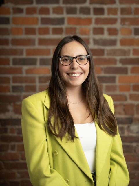 Professional headshot of a person with long dark hair wearing glasses, a bright green blazer, and a white top, standing in front of a brick wall and facing the camera.