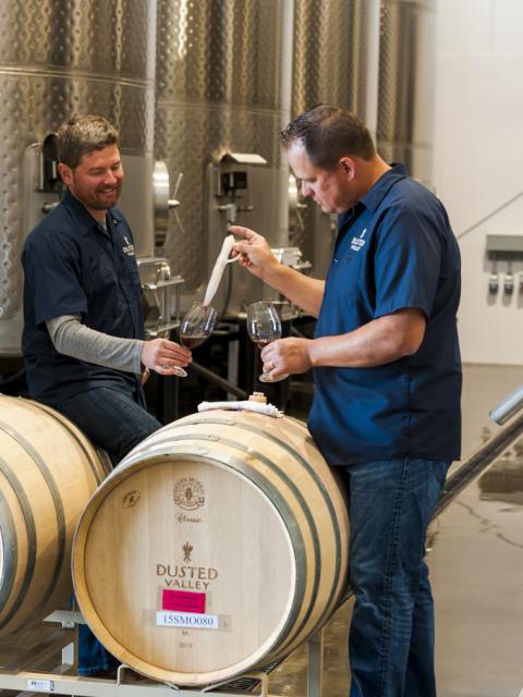 Two people in a winery cellar transferring wine from a barrel into glasses, with stainless steel tanks and wooden barrels behind them.