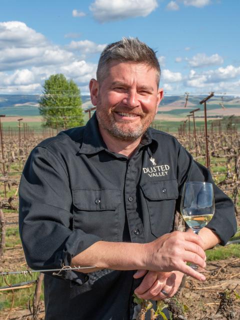 Person standing in a vineyard holding a glass of white wine, with rows of vines and rolling hills in the background.