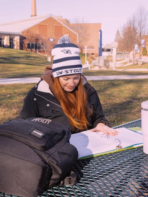 Student wearing a UW-Stout winter hat studies at an outdoor table on campus, with a backpack and binder open in front of them.