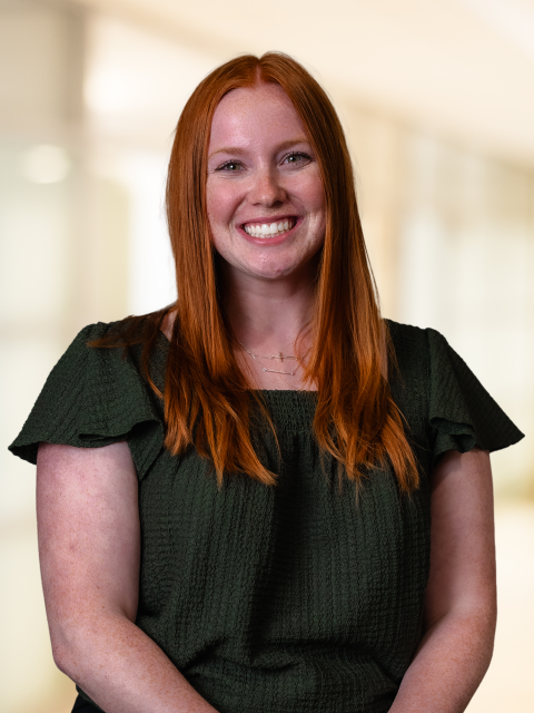 Person with long red hair smiling at the camera, seated against a softly blurred indoor background.