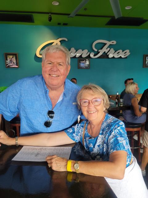 Two older adults seated at a restaurant table, smiling at the camera, with a turquoise wall and “Green Tree” sign behind them.