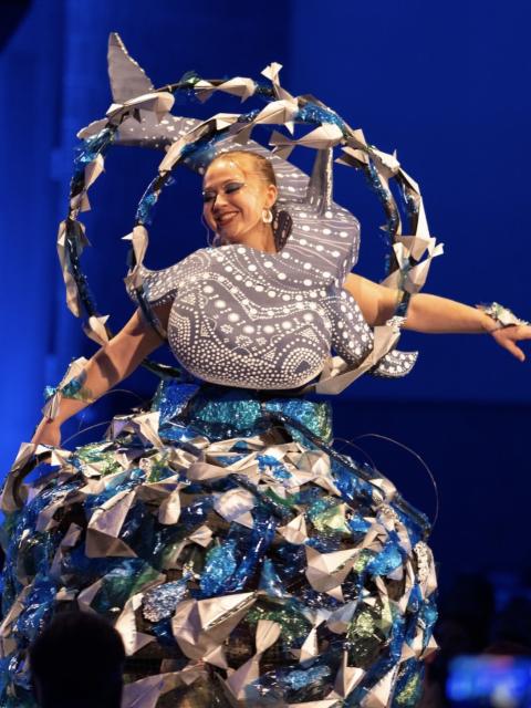 Model on stage wearing a large, ocean-themed sculptural dress with a whale shape and blue, white and metallic materials.