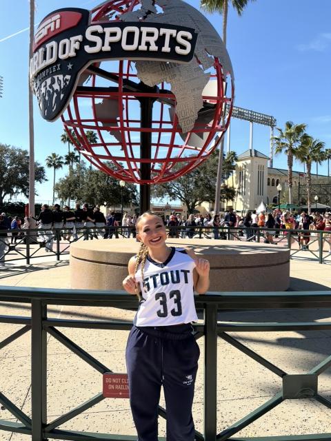 Person wearing a UW-Stout jersey stands in front of the ESPN Wide World of Sports globe at a busy outdoor venue.