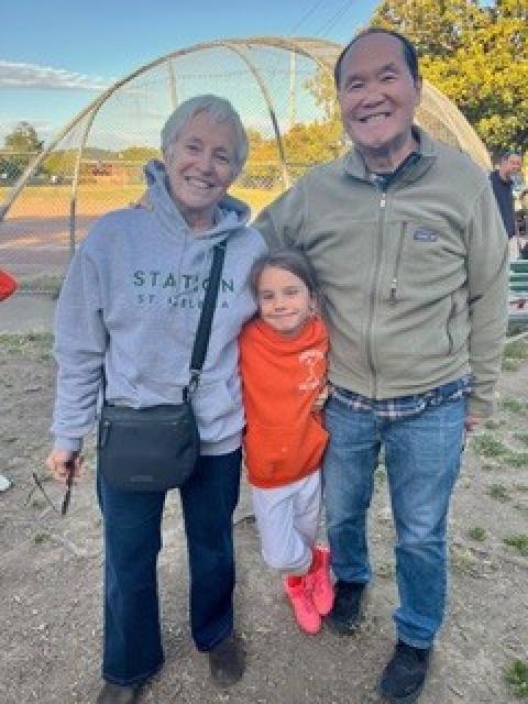 Three people posing together outdoors near a baseball field.