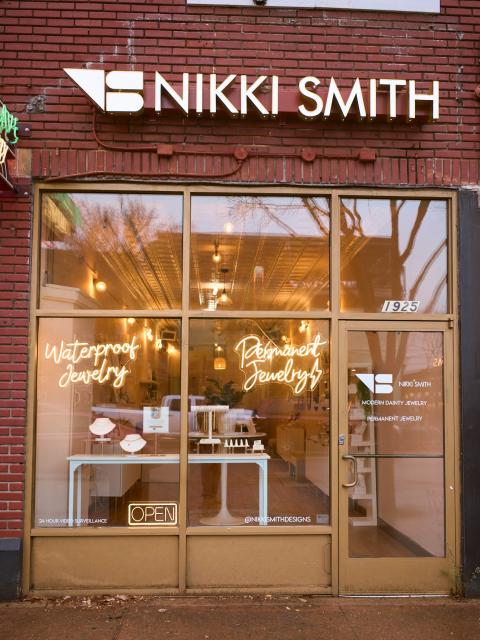 Exterior of a Nikki Smith jewelry storefront with large windows displaying jewelry and illuminated signs reading “Waterproof Jewelry” and “Permanent Jewelry.”
