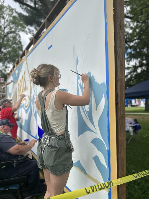 Person paints a light blue section of a large mural panel alongside other artists working nearby.