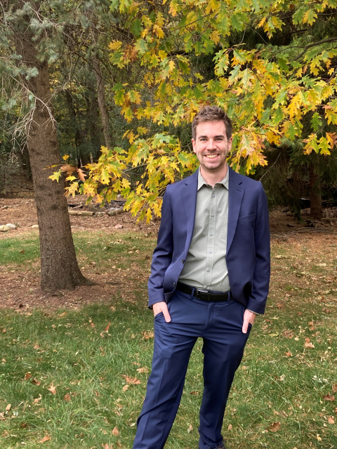 Michael Hilliger stands outdoors in a blue suit beneath a tree with yellow autumn leaves.