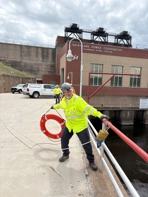 A utility worker wearing safety gear demonstrates a water rescue exercise near a hydroelectric facility while holding a life ring.
