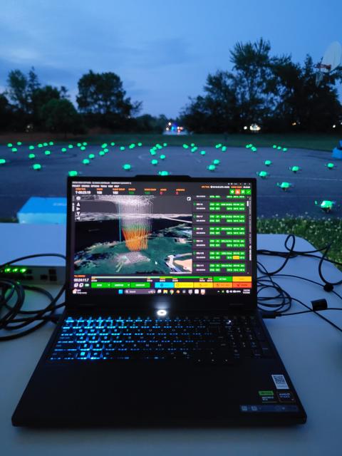 Laptop displaying drone fleet software beside dozens of illuminated drones arranged on a field at dusk.