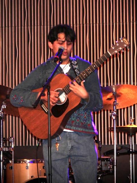 A student performs on stage, singing into a microphone while playing an acoustic guitar, with a drum set visible in the background.