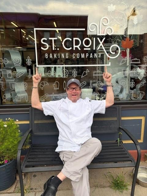 A person wearing a white chef coat, tan pants and a gray cap sits on a black bench outside St. Croix Baking Company with both arms raised and pointing toward the bakery sign in the window behind them.