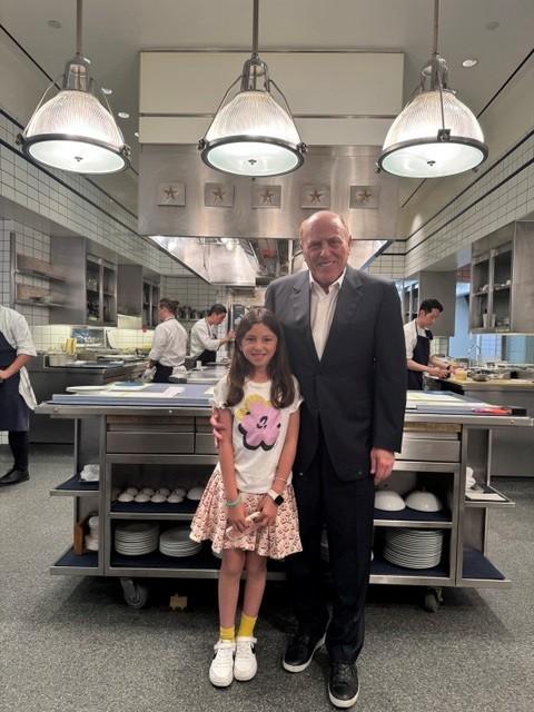 A person in a suit stands beside a child in a commercial kitchen, with chefs working in the background and stainless-steel equipment throughout the space.