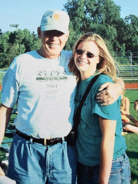 Two people stand together on an outdoor athletic field, with one person's arm around the other's shoulder. Trees and bleachers are visible in the background.
