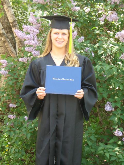 Brianne Maier wears graduation regalia and holds a UW-Stout diploma cover in front of blooming lilac bushes.
